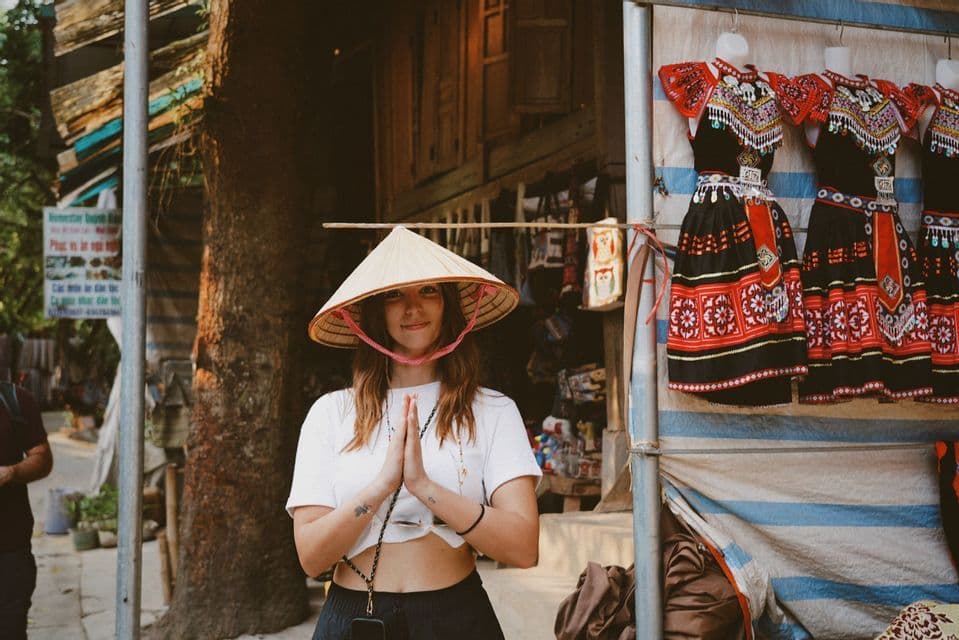 Une femme portant un chapeau conique sourit, les mains jointes, devant un étal de marché présentant des robes traditionnelles.