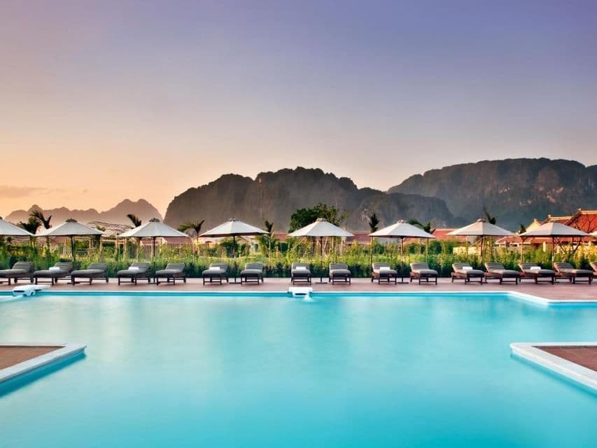 Una fila de tumbonas y sombrillas vacías bordea una gran piscina, con vistas a las montañas al atardecer.