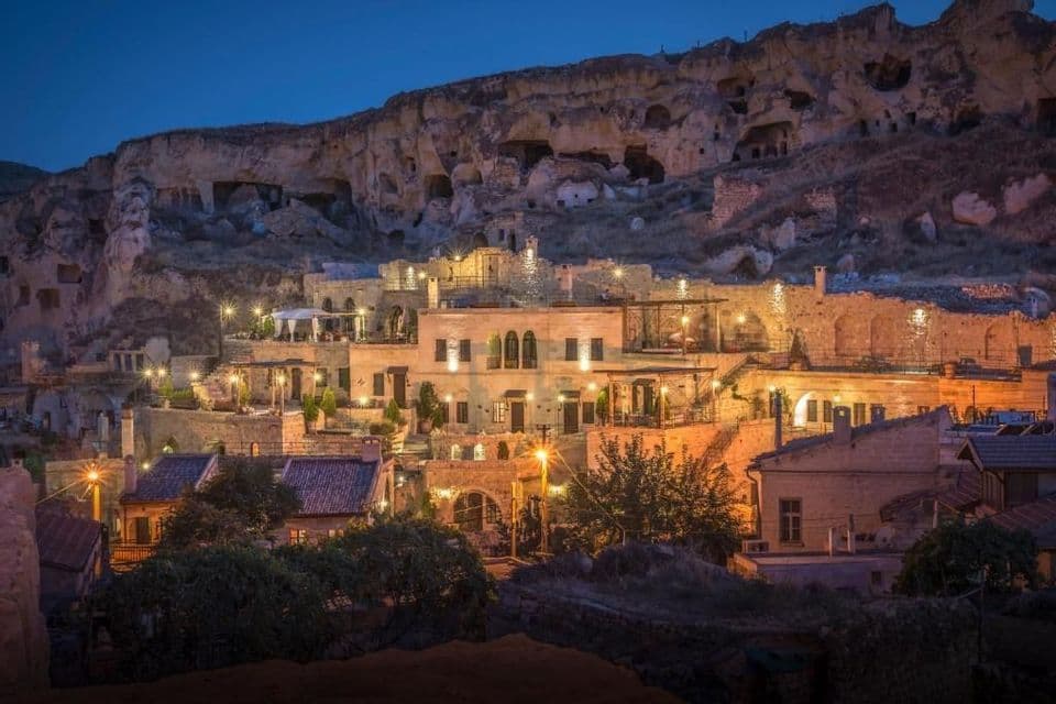 Edificios de piedra iluminados y viviendas rupestres excavadas en una ladera rocosa al anochecer.