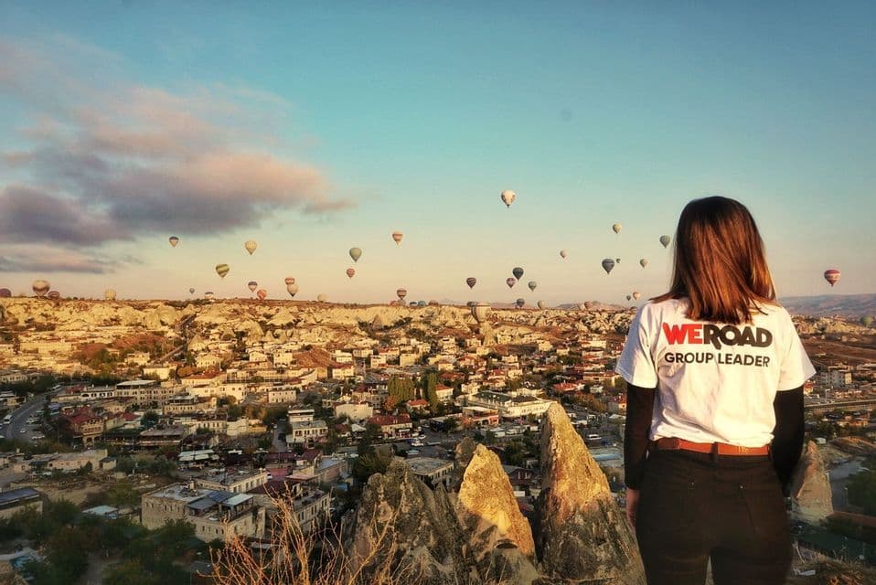Un líder de grupo de WeRoad está de pie en un acantilado con vistas a un pueblo mientras decenas de globos aerostáticos flotan en el cielo al amanecer.