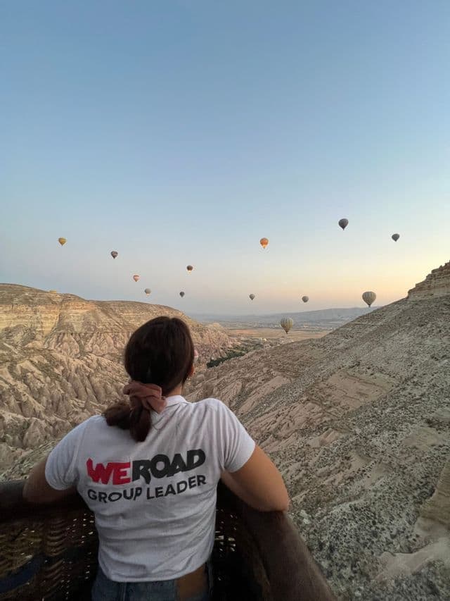 Un chef de groupe WeRoad observe depuis une nacelle tandis que des montgolfières dérivent au-dessus d'un paysage de canyon rocheux au lever du soleil.