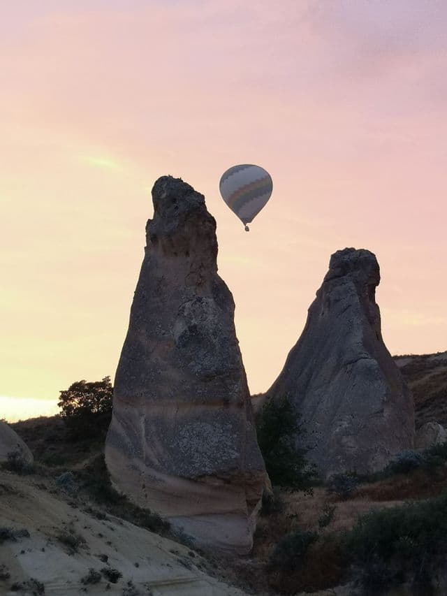 Un ballon à air chaud flotte au-dessus de formations rocheuses coniques sur un ciel rose au lever du soleil.