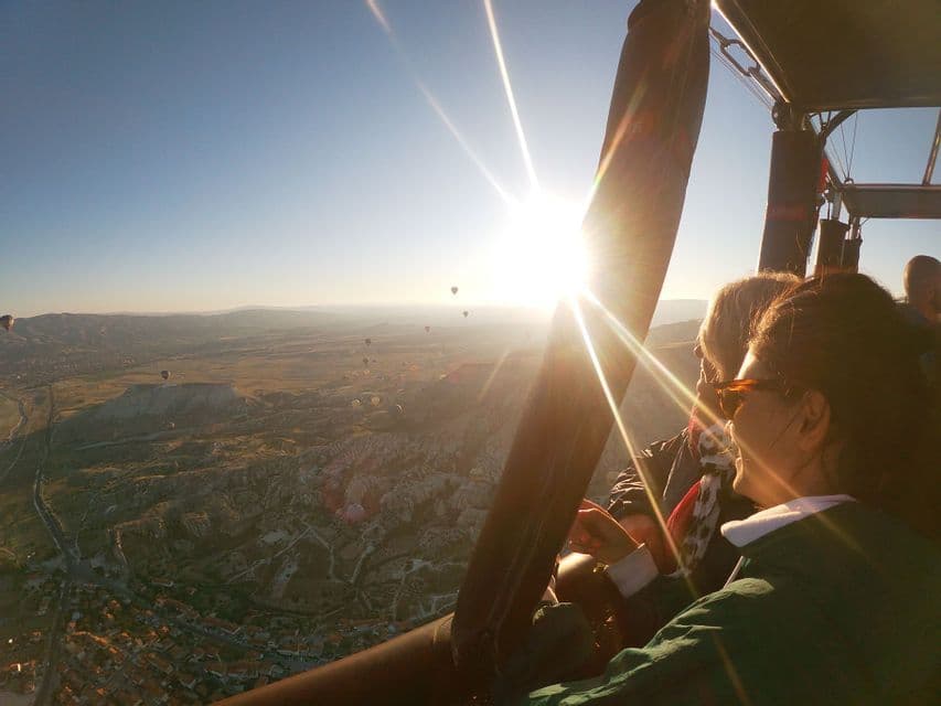 A WeRoad group trip watches the sunrise from a hot air balloon basket, looking out over a valley filled with other balloons.