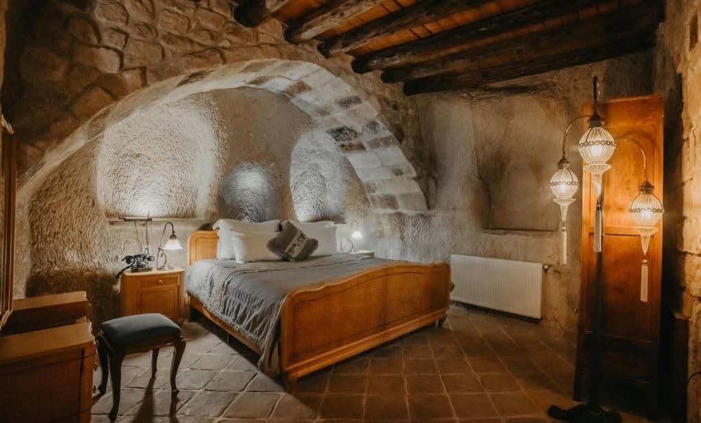 A warmly lit bedroom inside a stone cave, featuring a wooden bed under a vaulted ceiling with exposed wooden beams.