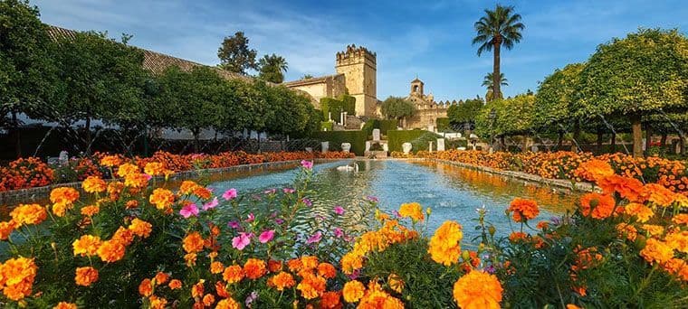 A long reflecting pool in a formal garden is lined with orange marigolds and trees, with a historic stone building in the background.