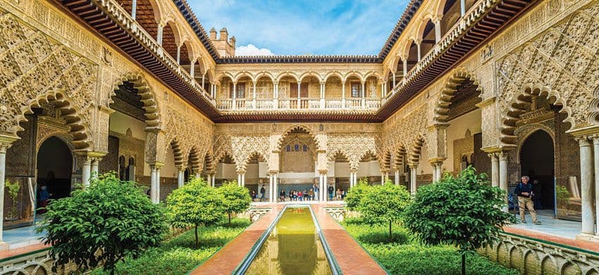 An ornate Moorish courtyard with a central reflection pool, gardens, and intricately carved archways under a blue sky.