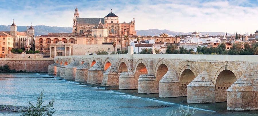 A historic stone bridge with many arches crosses a river towards a city with ancient buildings at sunset.