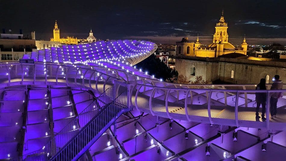 A modern architectural structure with a winding walkway, illuminated in purple at night, overlooking a city with historic buildings.
