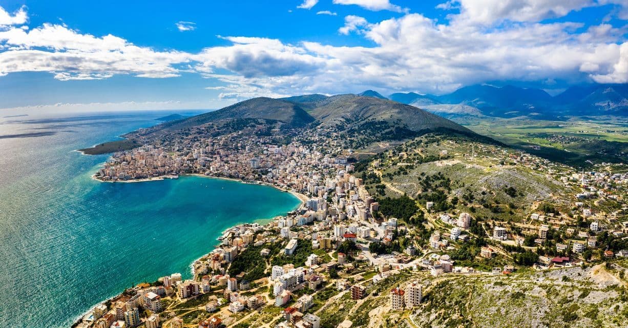 Aerial view of a coastal city curving around a turquoise bay, with green hills and mountains in the background.