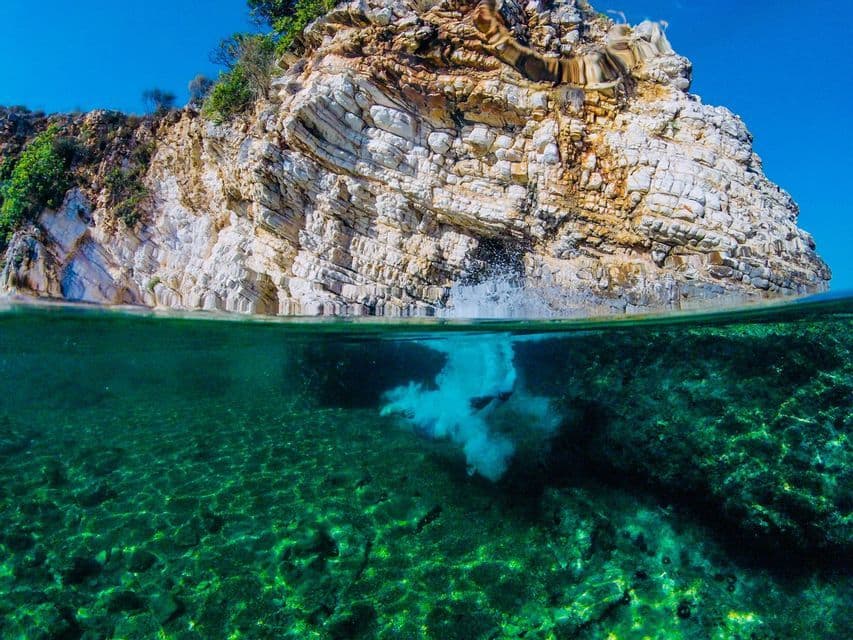 A split-view shot of a person diving from a rocky cliff into clear green water, with a splash on the surface and bubbles below.