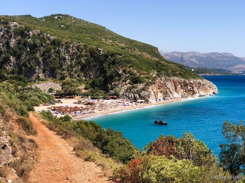 A view from a dirt path overlooking a crowded sandy beach with thatched umbrellas at the base of a green cliff and turquoise sea.