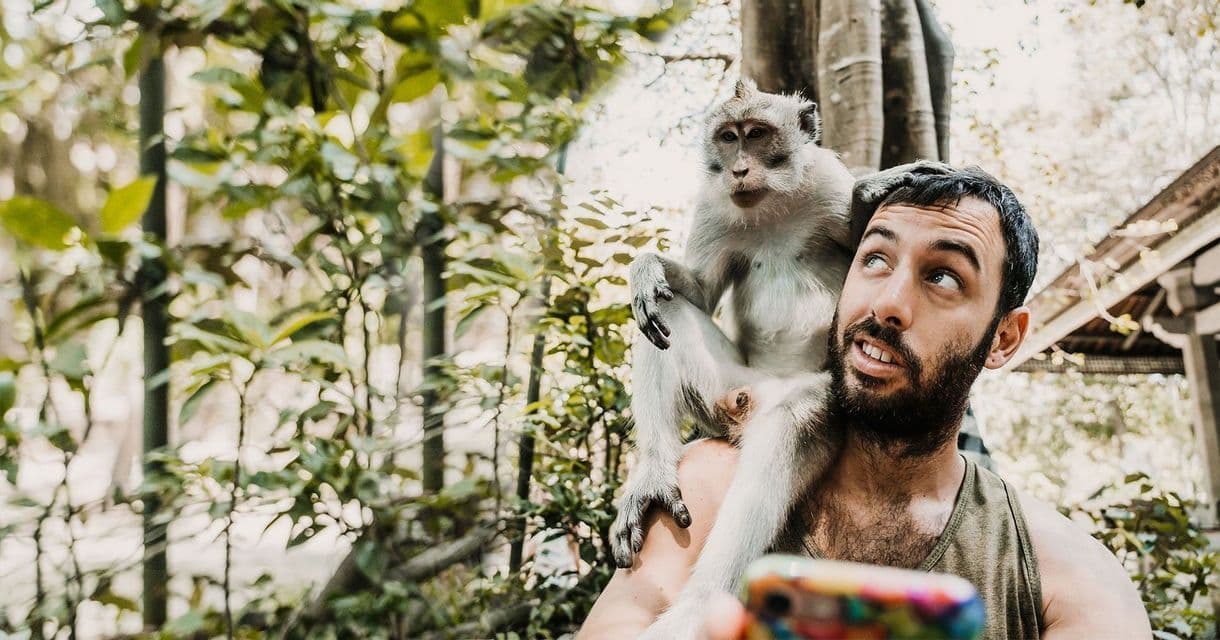 Una piccola scimmia si siede sulla spalla di un uomo barbuto mentre si scatta un selfie in una foresta verde.