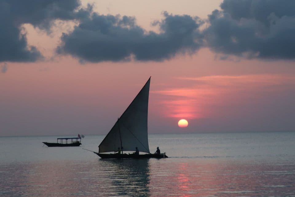 A sailboat with people on board glides across calm water at sunset, with the sun low on the horizon under a pink and cloudy sky.
