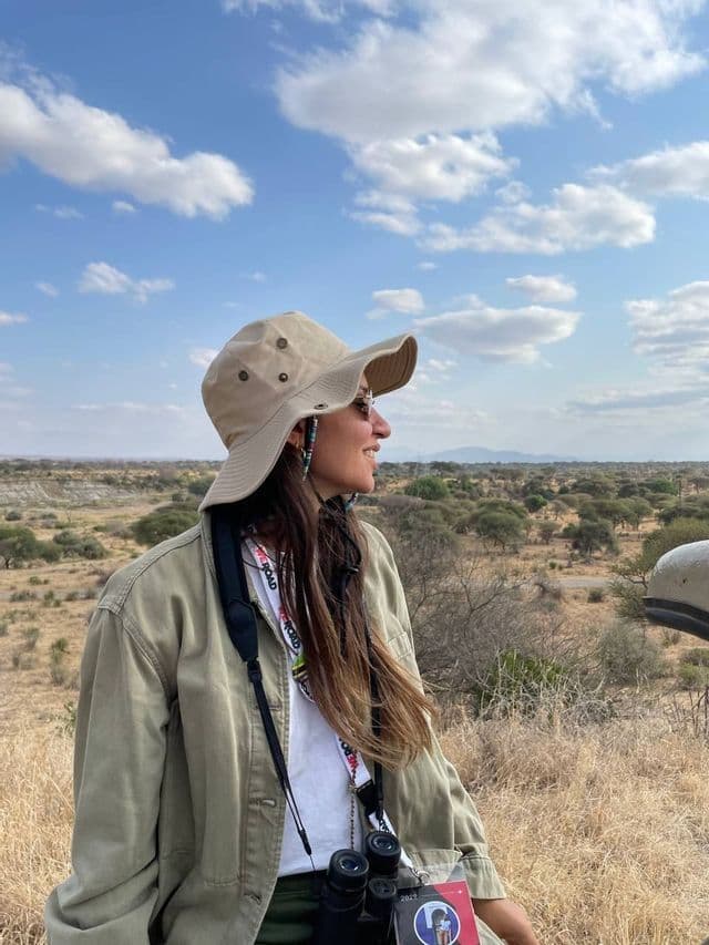 Une femme lors d'un voyage de groupe WeRoad, portant un chapeau de safari et des jumelles, contemple un paysage de savane.