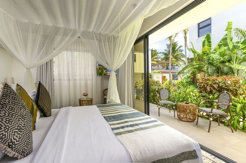 Une chambre lumineuse avec un lit à baldaquin blanc donne sur un patio privé avec des chaises et des plantes tropicales luxuriantes.