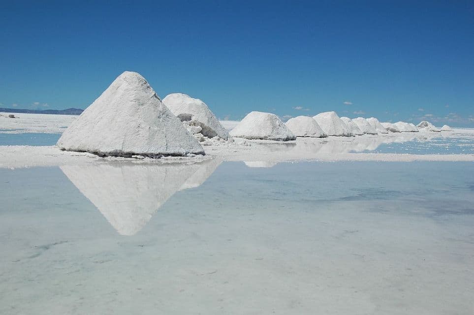 Una hilera de montículos de sal cónicos en una salina inundada se refleja en el agua tranquila bajo un cielo azul claro.