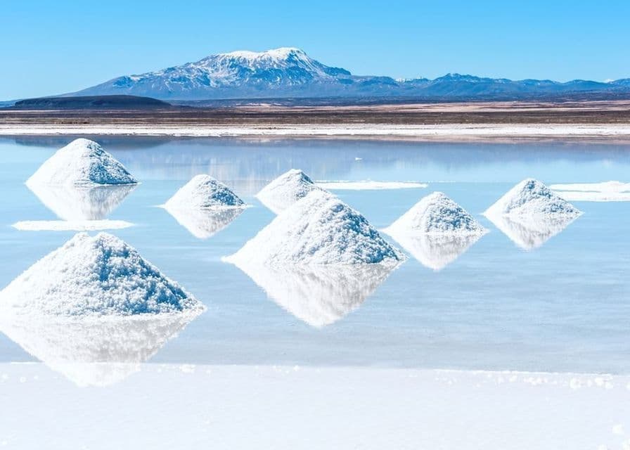 Cumuli di sale bianco giacciono in acqua calma su una salina, con montagne innevate sotto un cielo azzurro e limpido.