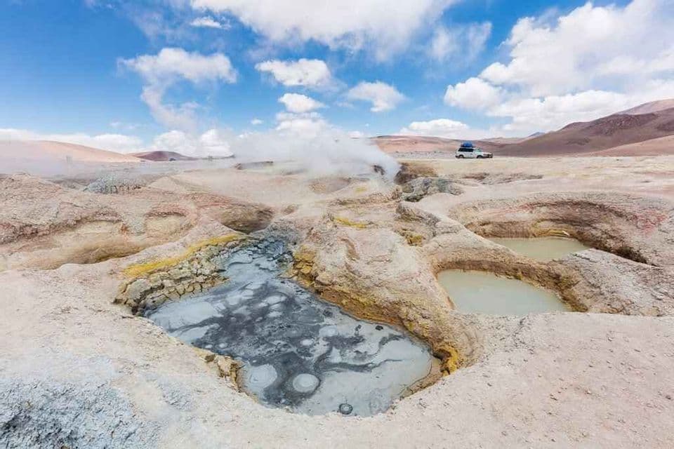 Ollas de lodo burbujeante y fumarolas humeantes en un vasto campo geotérmico bajo un cielo azul y nublado, con un vehículo a la distancia.