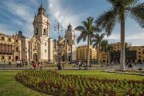 Una catedral histórica con torres gemelas domina una soleada plaza pública con palmeras, macizos de flores y gente paseando.