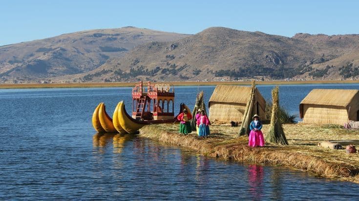 Des personnes en vêtements traditionnels colorés se tiennent sur une île flottante en roseaux, près de huttes au toit de chaume et d'un grand bateau en roseaux sur un lac.