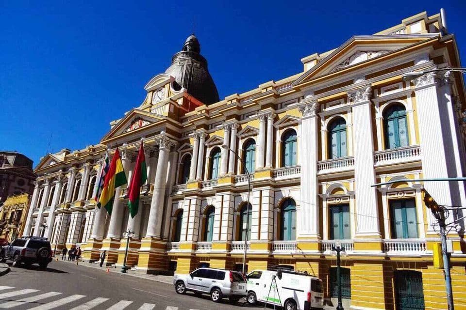Un gran edificio neoclásico amarillo y blanco con altas columnas, una cúpula y varias banderas se alza en una calle de la ciudad bajo un cielo azul claro.