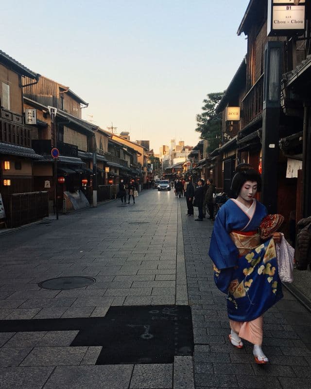 Une femme vêtue d'un kimono bleu traditionnel et maquillée de blanc marche le long d'une rue pavée bordée de bâtiments traditionnels en bois au crépuscule.
