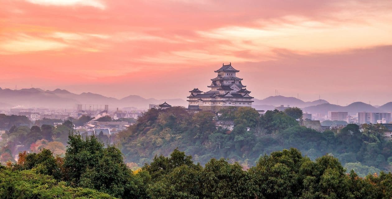 Eine traditionelle japanische Burg auf einem bewaldeten Hügel, die eine Stadt mit nebligen Bergen bei Sonnenuntergang überblickt.