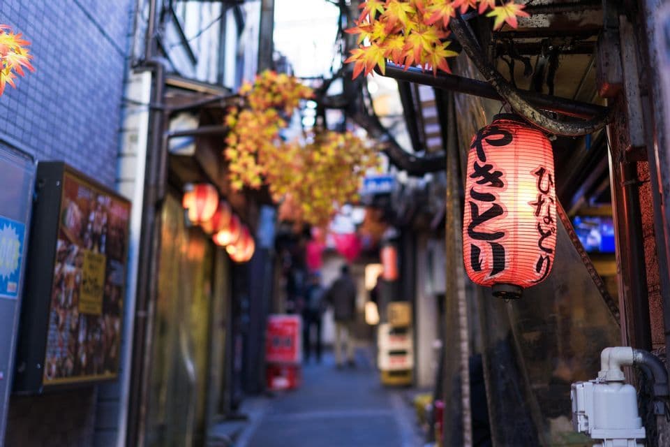 Nahaufnahme einer beleuchteten roten japanischen Papierlaterne mit Kalligraphie, die in einer engen Stadtgasse hängt, geschmückt mit Herbstblättern.