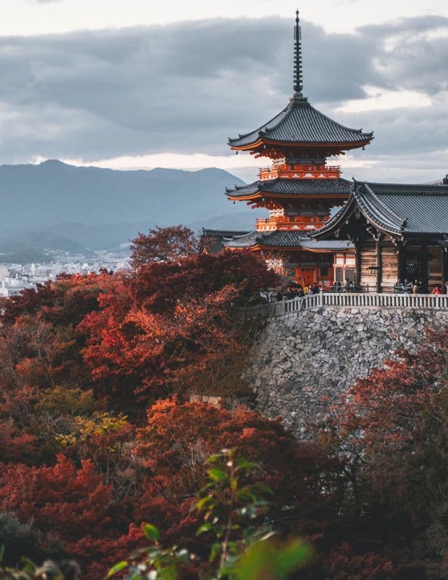 Une pagode japonaise rouge et noire surplombe une forêt dense au feuillage d'automne rouge, avec des montagnes en arrière-plan sous un ciel nuageux.