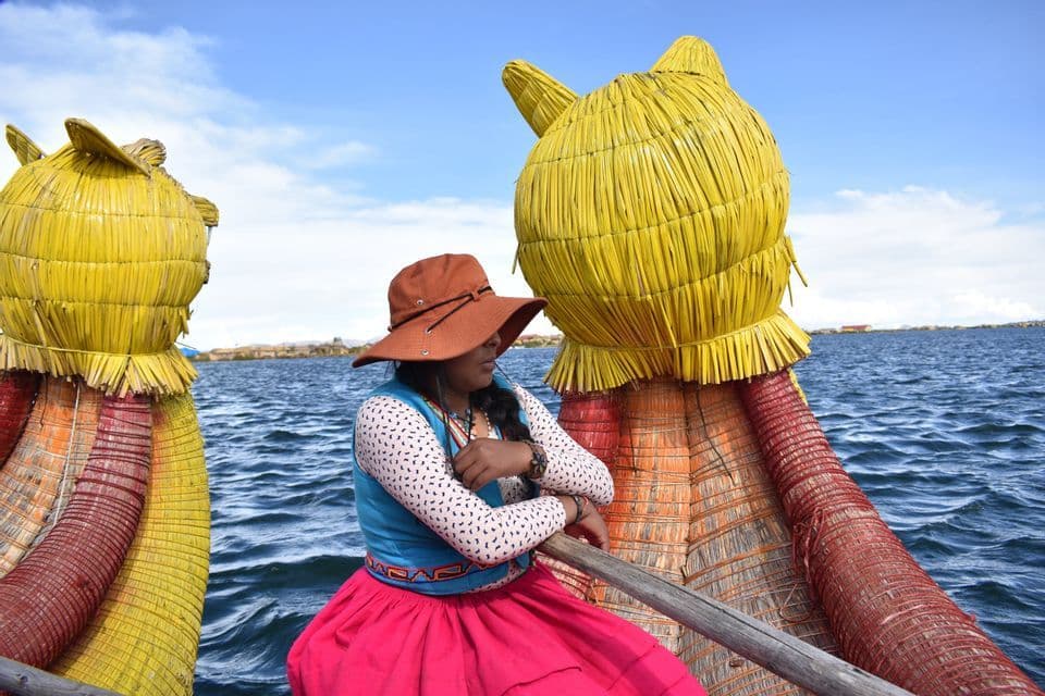 Eine Frau in farbenfroher Tracht sitzt in einem dekorativen Schilfboot auf einem großen See unter einem teilweise bewölkten Himmel.