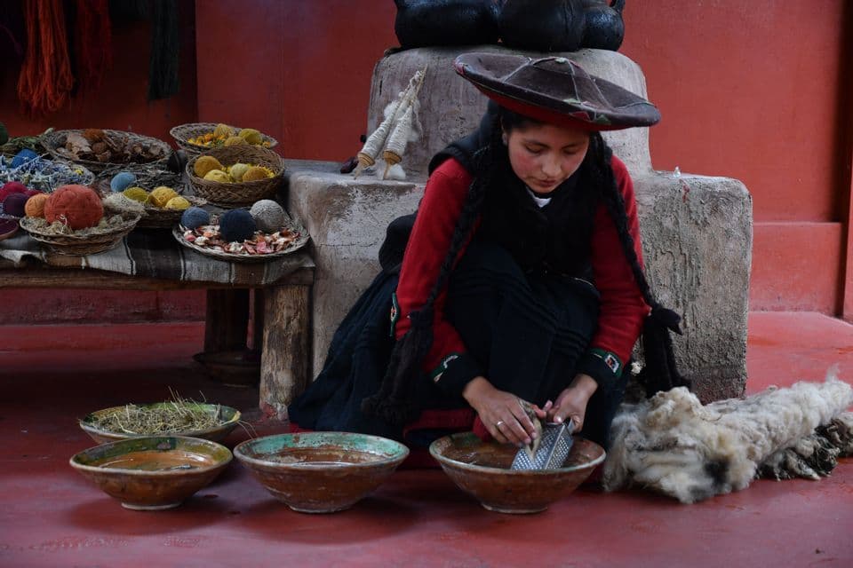 Une femme en tenue traditionnelle râpe une racine dans un bol pour fabriquer de la teinture naturelle, entourée de laine brute et de fils colorés.