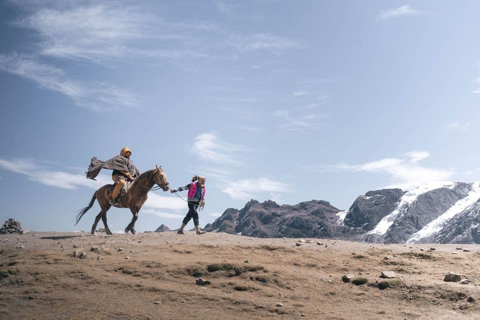 Une participante d'un voyage de groupe WeRoad guide un cheval, accompagnée d'un guide en poncho, à travers un paysage rocheux de haute altitude avec des montagnes enneigées.