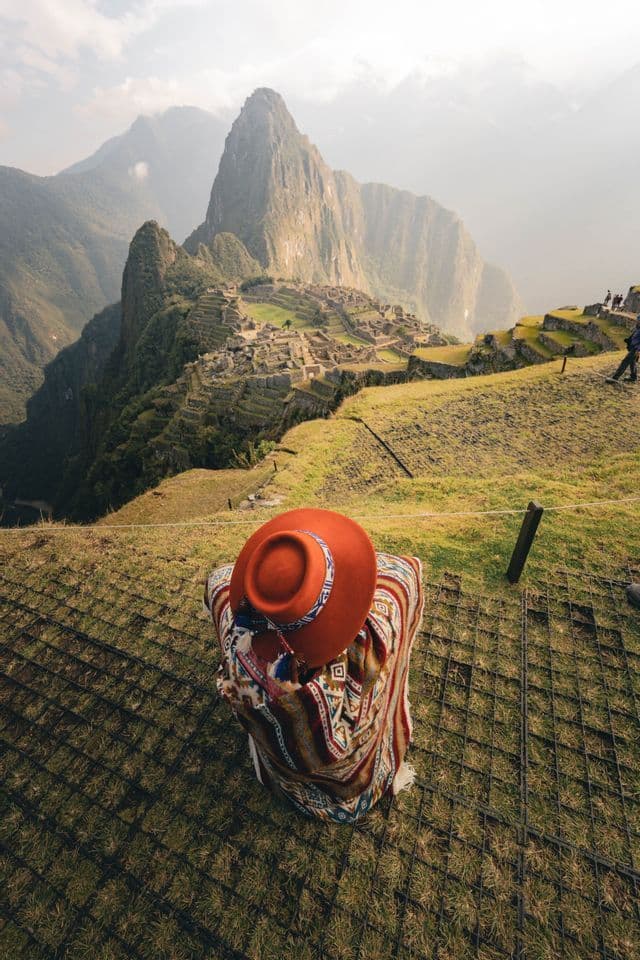 Una persona con un poncho colorato e un cappello arancione è seduta su una collina erbosa che domina antiche rovine e montagne.