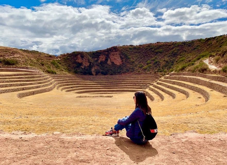Una donna con uno zaino siede su una sporgenza guardando grandi terrazzamenti agricoli concentrici in una valle sotto un cielo parzialmente nuvoloso.