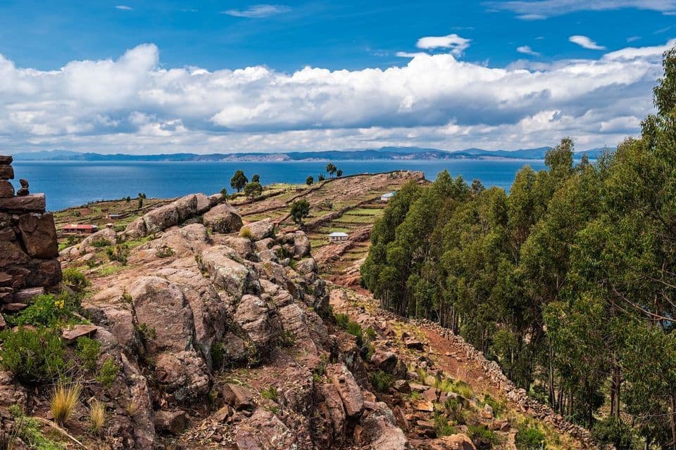 Une colline rocheuse avec des champs en terrasses et des arbres surplombe une grande étendue d'eau bleue sous un ciel partiellement nuageux.