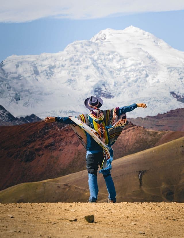 Una persona de espaldas vistiendo un colorido poncho y sombrero se para con los brazos abiertos ante una enorme montaña nevada.