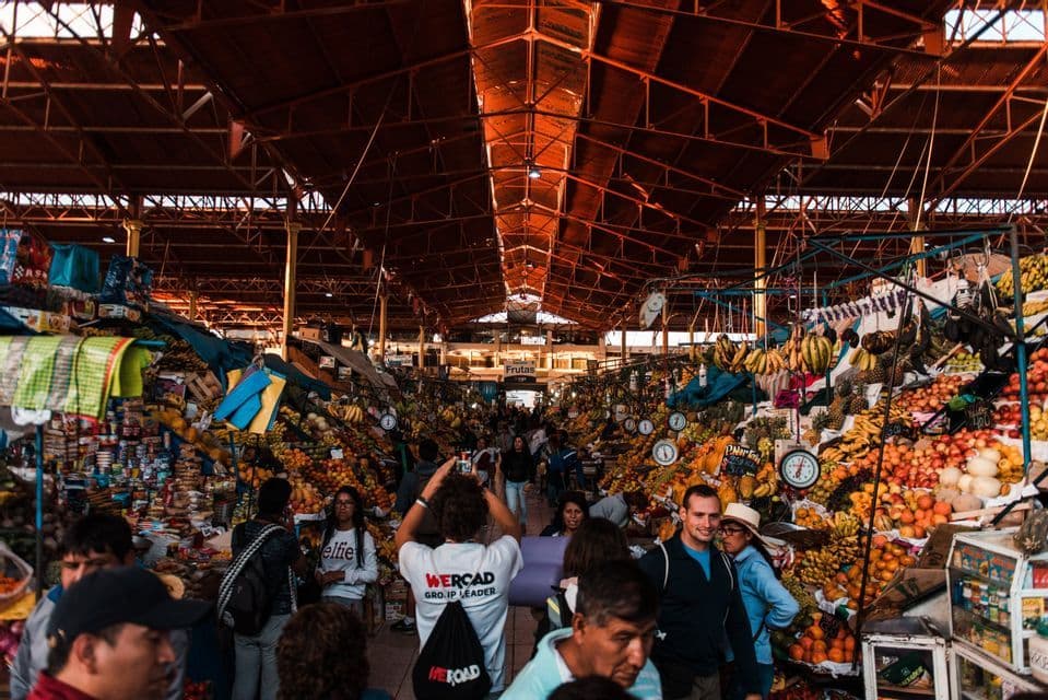 Un voyage de groupe WeRoad explorant un marché aux fruits intérieur animé avec un haut plafond à poutres métalliques.