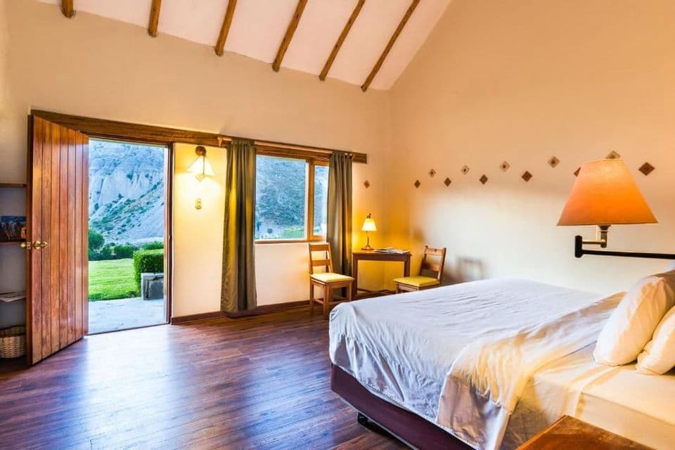 Ein Hotelzimmer mit gemachtem Bett und Holzboden hat offene Türen, die einen Blick auf eine Rasenfläche und felsige Berge freigeben.