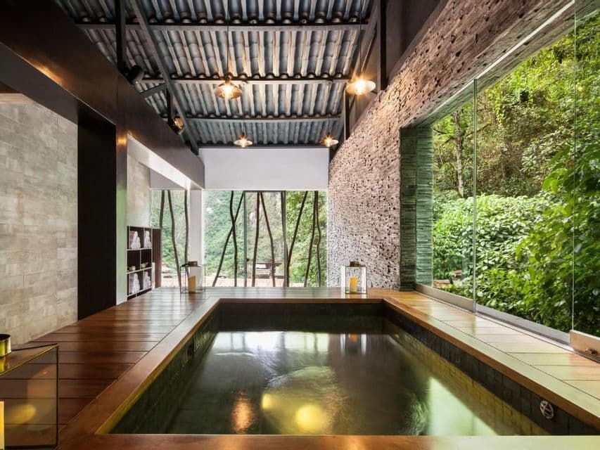 Une piscine intérieure dans une pièce avec une terrasse en bois, un mur en pierres empilées et de grandes fenêtres avec vue sur une forêt luxuriante.