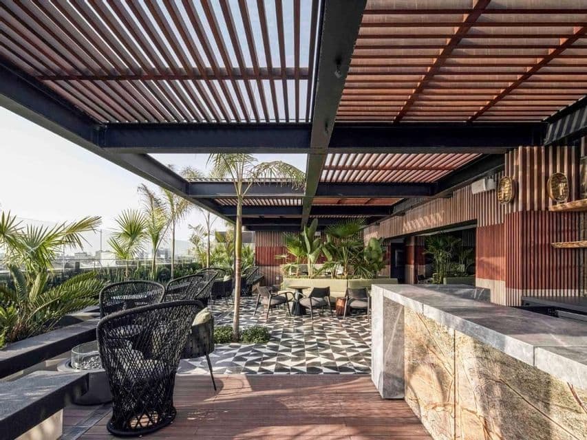 Lounge de toit moderne avec pergola en bois, bar en pierre, chaises tressées noires et sol carrelé à motifs, entouré de plantes.