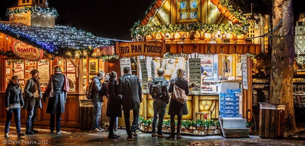 Eine Gruppe von Menschen steht nachts vor hell beleuchteten und dekorierten Essensständen auf einem Außenmarkt.