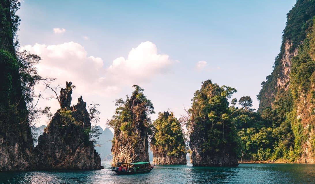 Un viaje en grupo de WeRoad en barco navega entre grandes acantilados de piedra caliza cubiertos de árboles que se elevan desde aguas azules y tranquilas.