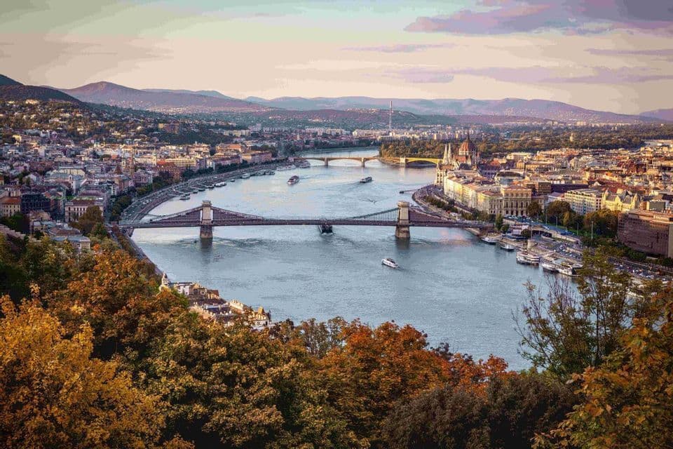 Una vista aérea de una ciudad con un río ancho, múltiples puentes y barcos, vista sobre coloridos árboles de otoño con colinas al fondo.