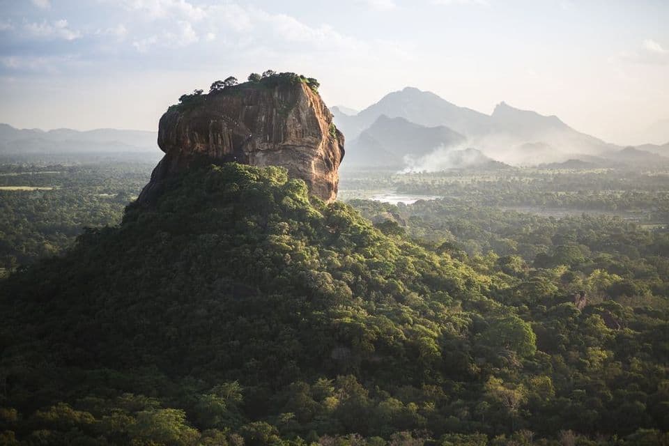 A large rock monolith rises from a dense green forest, with a hazy mountain range visible in the background.