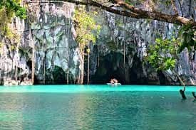 A small boat with people paddles into a large cave entrance from a turquoise lagoon surrounded by lush green cliffs.