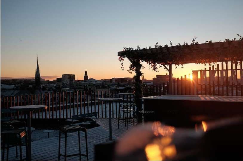 Une terrasse de toit vide avec des tables et des tabourets surplombe un paysage urbain orné d'un clocher d'église, baigné par un coucher de soleil éclatant.