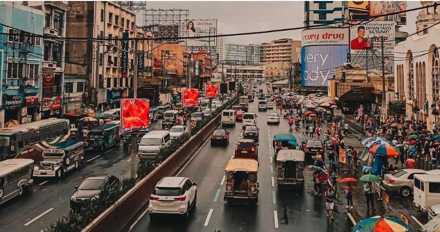 A high-angle view of a congested city street with cars, jeepneys, and buses on a wet road, with pedestrians walking on the sidewalks.