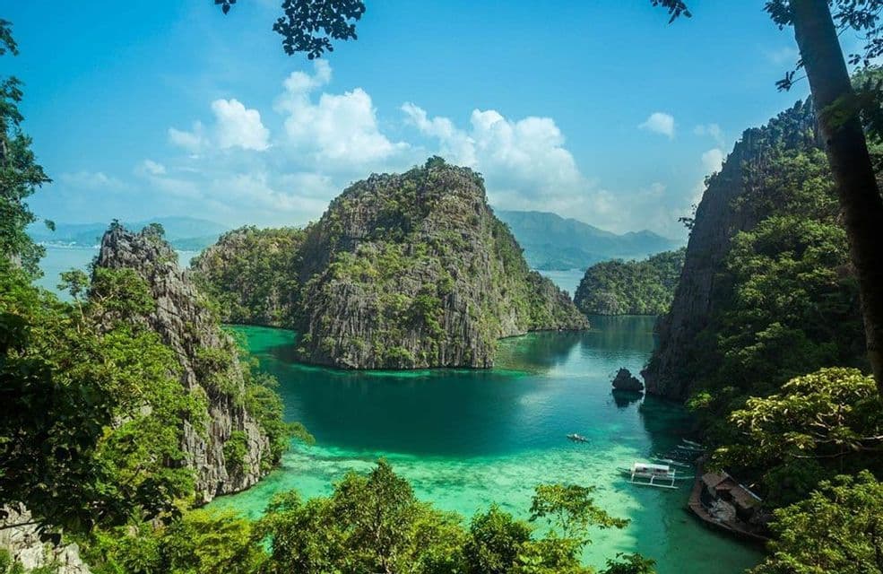 A view from above shows a sunken shipwreck and a boat in a clear turquoise lagoon, surrounded by lush, rocky islands under a blue sky.