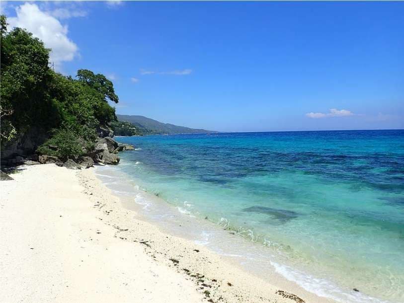 A pristine white sand beach meets clear, turquoise ocean waters next to a rocky, tree-covered shoreline under a bright blue sky.