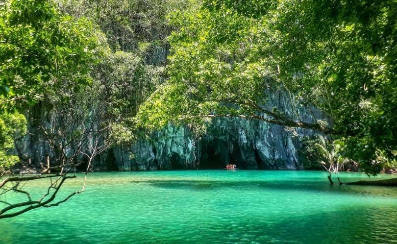 A WeRoad group trip in a boat sails on turquoise water towards a cave opening in a large, jungle-covered rock cliff.